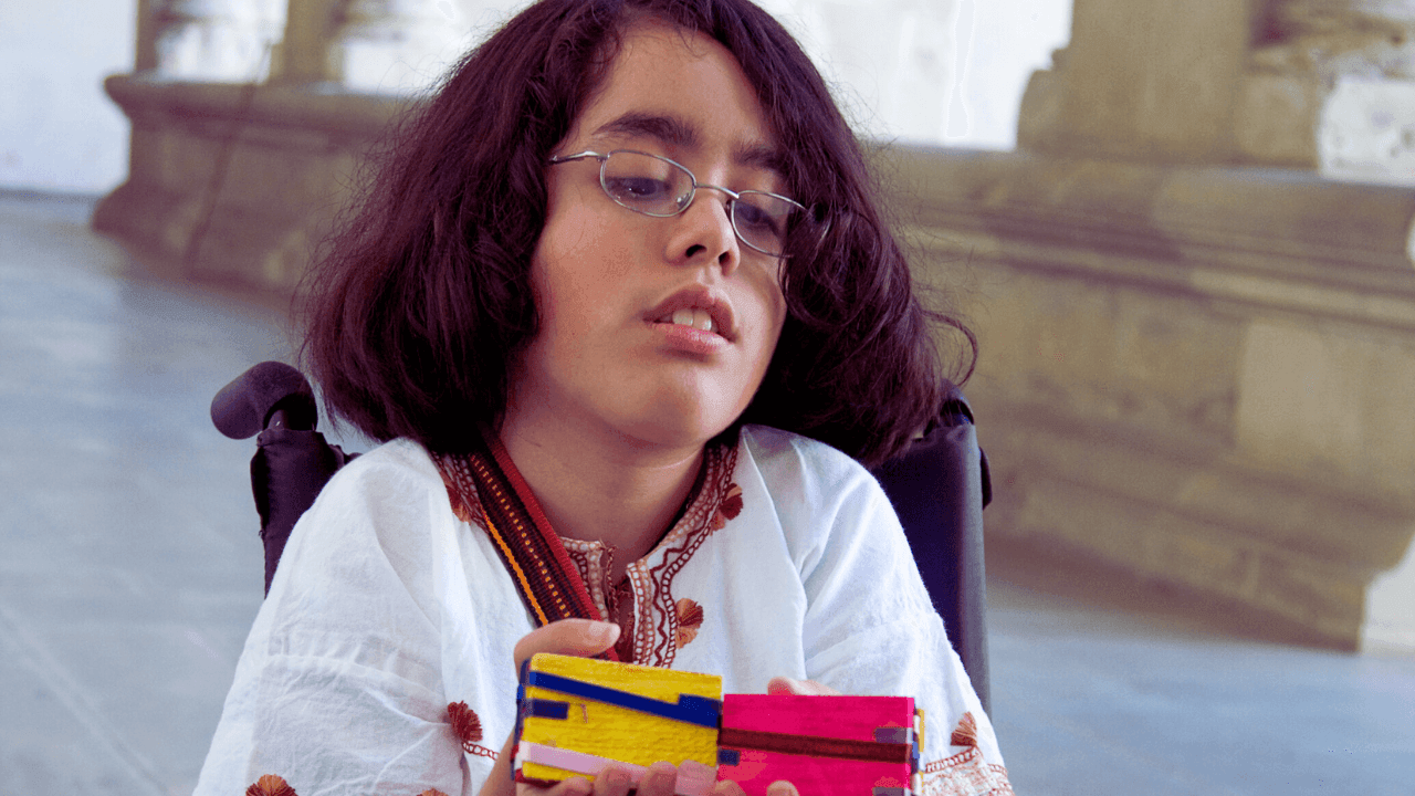 Young person in a wheelchair holding colorful objects, wearing glasses and a white embroidered shirt.