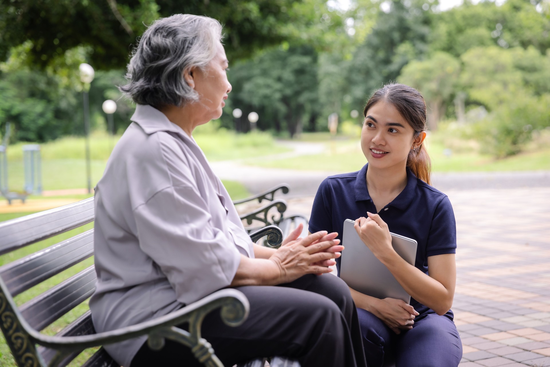Young caregiver discussing senior care plans with an elderly woman using a laptop while sitting on a park bench, emphasizing attentive support and personalized elderly care in a serene environment.
