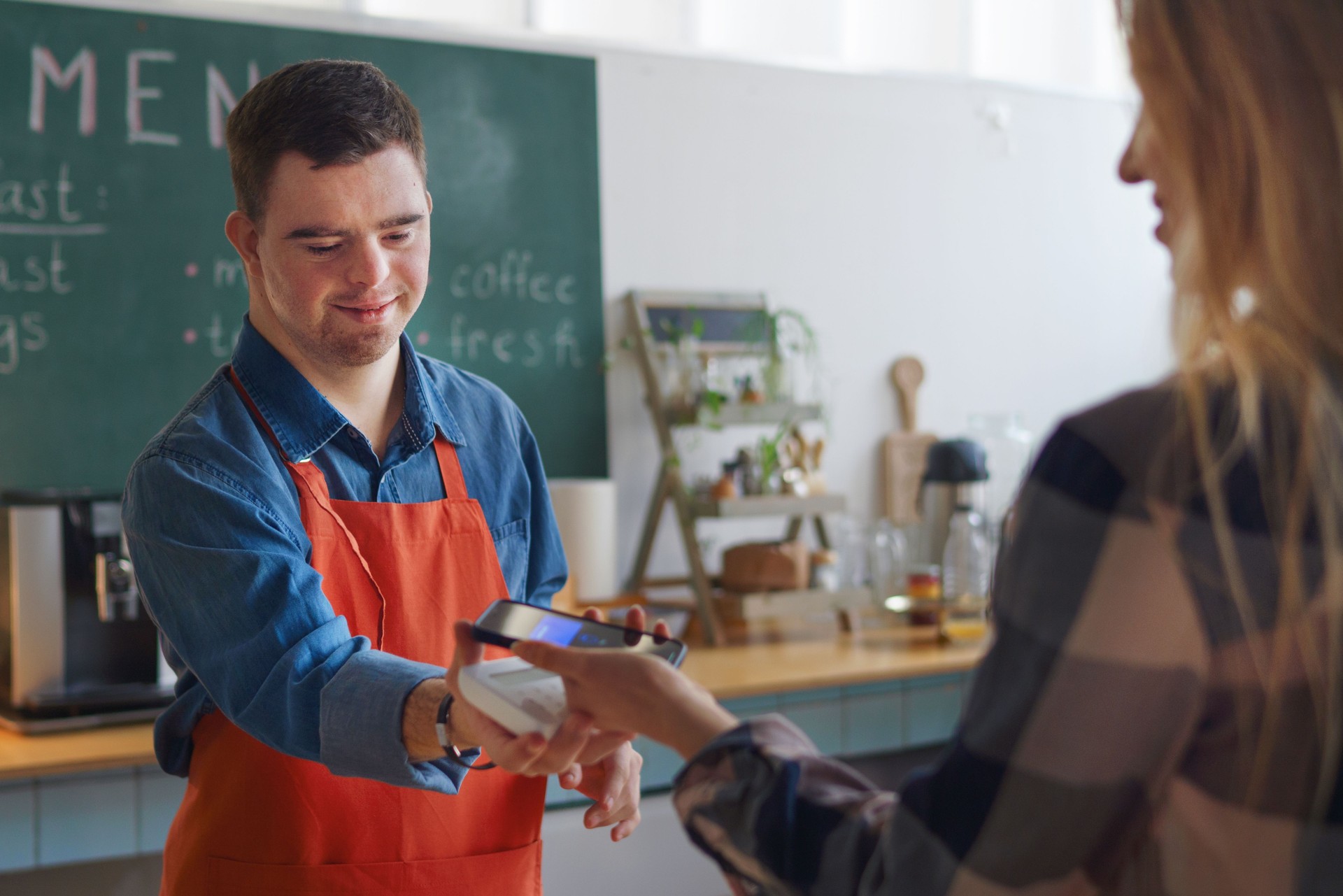 Cheerful young Down Syndrome waiter taking contactless smartphone payment from costumer in take away restaurant.
