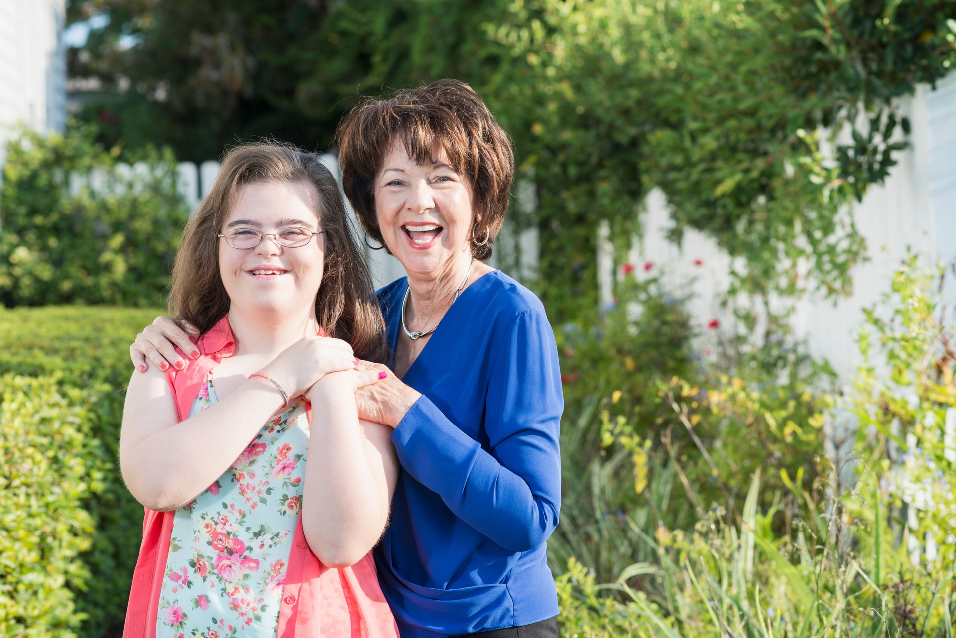 Aging woman standing beside her adult daughter with Down syndrome in a garden near a white picket fence—representing future planning, caregiver transition, and disability support for aging parents