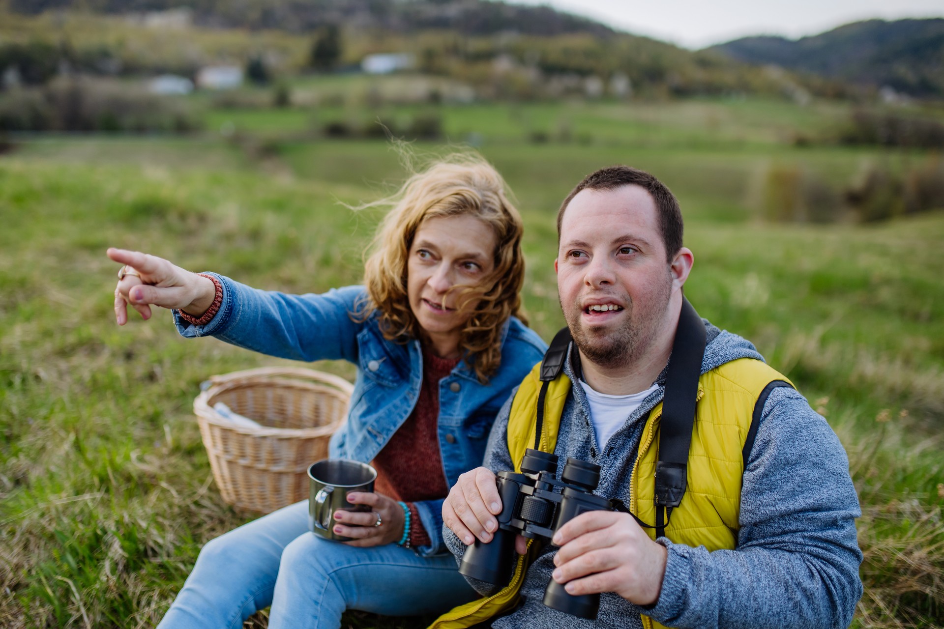 Happy young man with Down syndrome sitting in nature with his sister, looking at the view through binoculars—symbolizing sibling caregiving, disability support, and future planning for adults with developmental disabilities