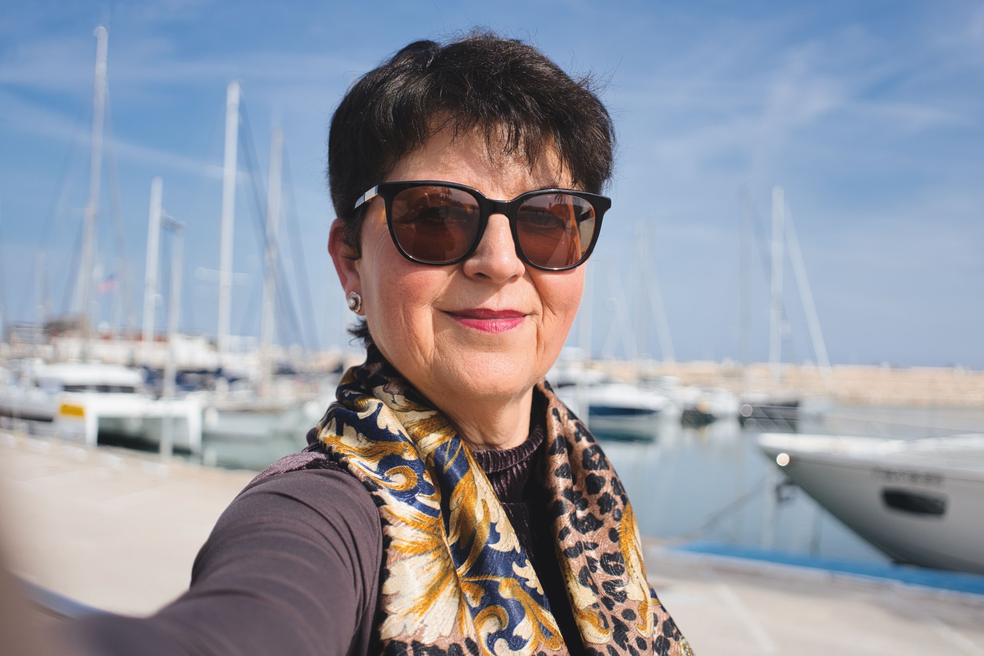 Stylish mature woman with sunglasses and a colorful scarf takes a selfie at a marina full of sailboats on a beautiful sunny day, enjoying her leisure time