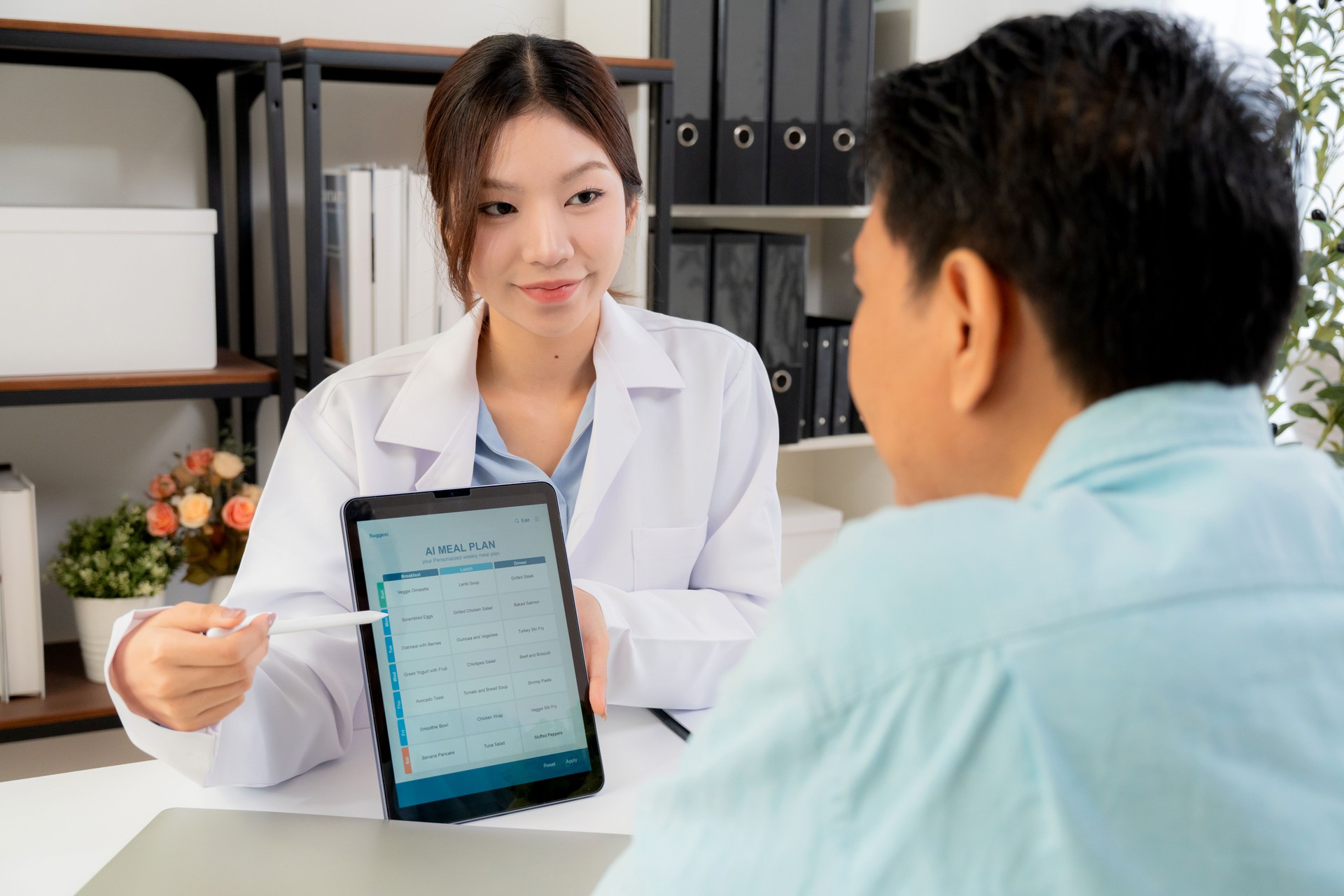 Asian nutritionist using tablet to explaining personalized meal plan with AI to patient during healthcare consultation, healthy lifestyle, woman dietitian showing nutrition plan with AI on tablet.