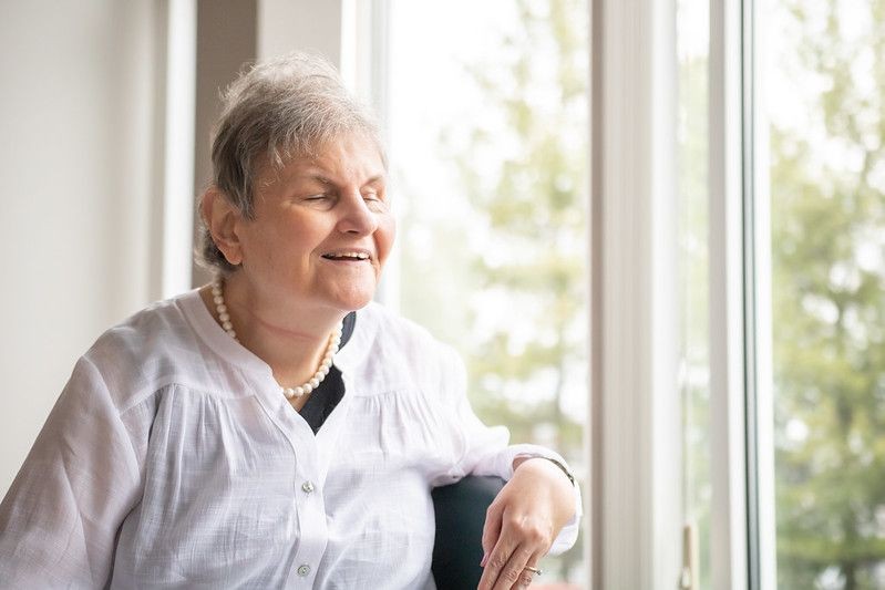 Aging blind woman wearing a white blouse and pearls, seated in front of a window with a nature view—representing aging with disabilities, dignified care, and future planning support for adults with vision impairments